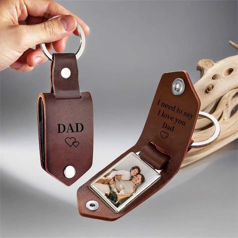 May include: A brown leather keychain with a silver key ring. The keychain is engraved with the word "DAD" and two hearts. The keychain opens to reveal a photo. The inside of the keychain is engraved with the words "I need to say I love you Dad".