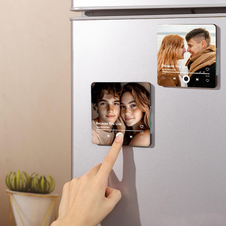 May include: Two square magnets with photos of couples are displayed on a silver refrigerator. One magnet shows a couple with the text "Because You Live" and a music player interface. The other magnet shows a couple with the same text and interface.