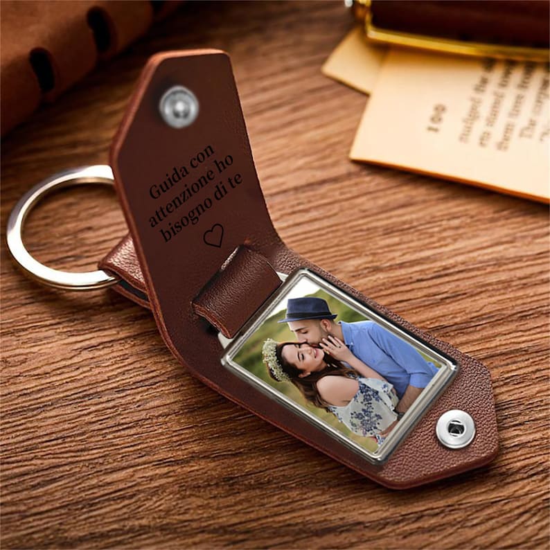 May include: A brown leather keychain with a silver key ring. The keychain has a rectangular photo of a couple and the engraved text "Guida con attenzione ho bisogno di te" with a heart symbol.