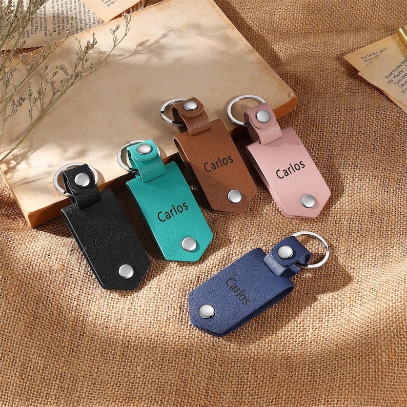 May include: A collection of leather keychains in various colors: black, teal, brown, pink, and blue. Each keychain features a silver-tone metal ring and a snap closure. The name "Carlos" is printed on each keychain.