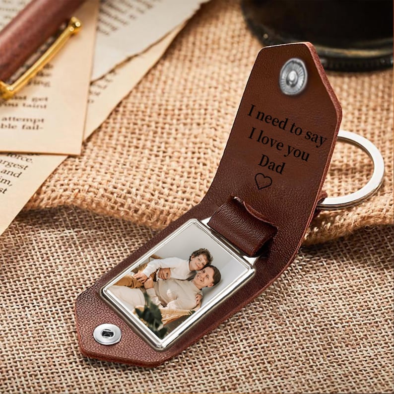 May include: A brown leather keychain with a silver photo frame. The frame contains a photo of a man and a child. The keychain has the engraved text "I need to say I love you Dad" with a heart symbol.