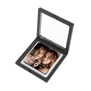 May include: A black shadow box with a clear glass front, displaying a photo of two people. The photo has a music player interface overlay with the text "Because You Live". The shadow box is open, revealing the photo.