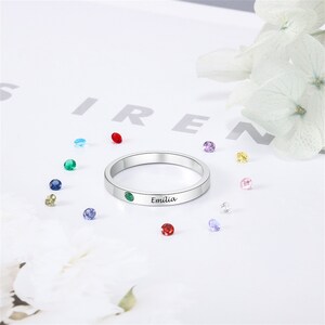 May include: A silver ring with a green gemstone and the name "Emilia" engraved on the band. The ring is surrounded by colorful gemstones.