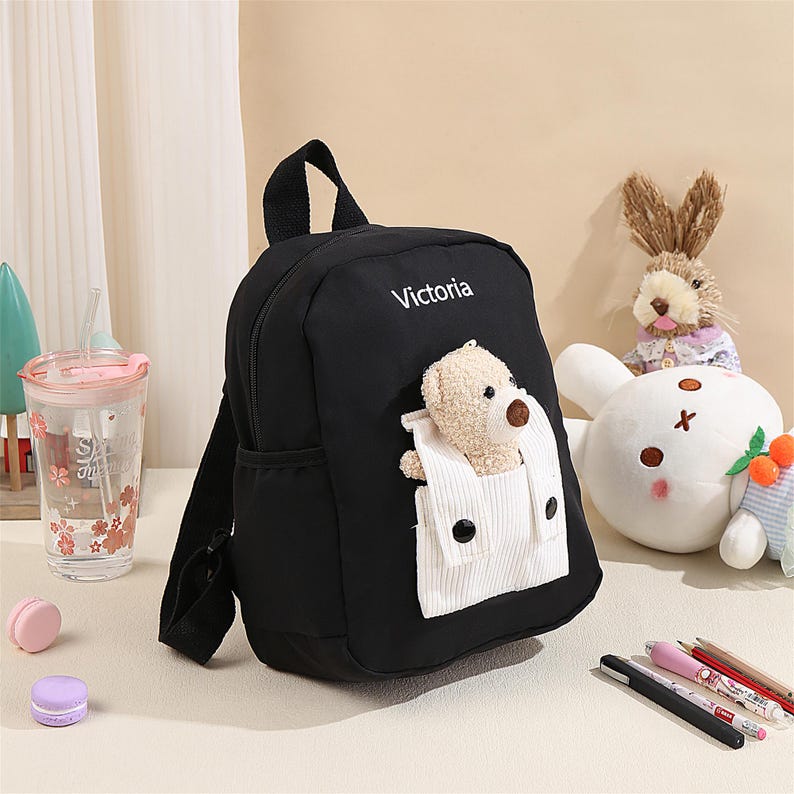 May include: A black backpack featuring the name Victoria printed in white text. The front includes a white corduroy pocket containing a small, beige plush teddy bear with a brown nose. The bag has black straps and a top handle for carrying.