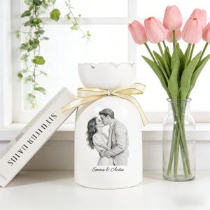 Puede incluir: Jarr&oacute;n de cer&aacute;mica blanca con la parte superior festoneada, atado con una cinta crema. El jarr&oacute;n presenta una imagen en blanco y negro de una pareja bes&aacute;ndose, con los nombres "Emma & Acilia" debajo. Un ramo de tulipanes rosas en un jarr&oacute;n de cristal transparente est&aacute; al lado.