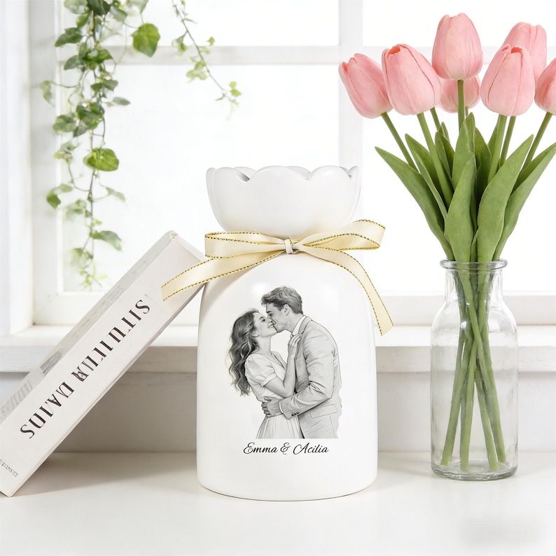 Puede incluir: Jarr&oacute;n de cer&aacute;mica blanca con la parte superior festoneada, atado con una cinta crema. El jarr&oacute;n presenta una imagen en blanco y negro de una pareja bes&aacute;ndose, con los nombres "Emma & Acilia" debajo. Un ramo de tulipanes rosas en un jarr&oacute;n de cristal transparente est&aacute; al lado.