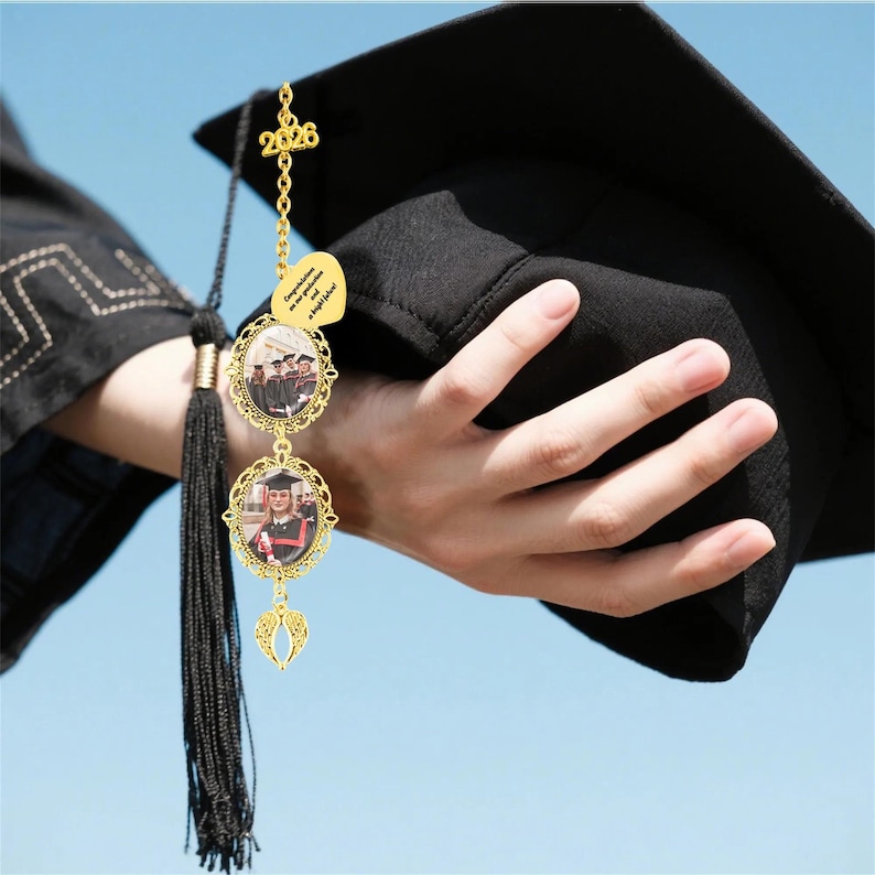 Pode incluir: Um adorno de formatura dourado com duas molduras ovais para fotos e uma pequena etiqueta em forma de cora&ccedil;&atilde;o. O adorno &eacute; preso a um borla preta e a um chap&eacute;u de formatura. A etiqueta diz "Parab&eacute;ns pela sua formatura e um futuro brilhante!" e o ano "2026".