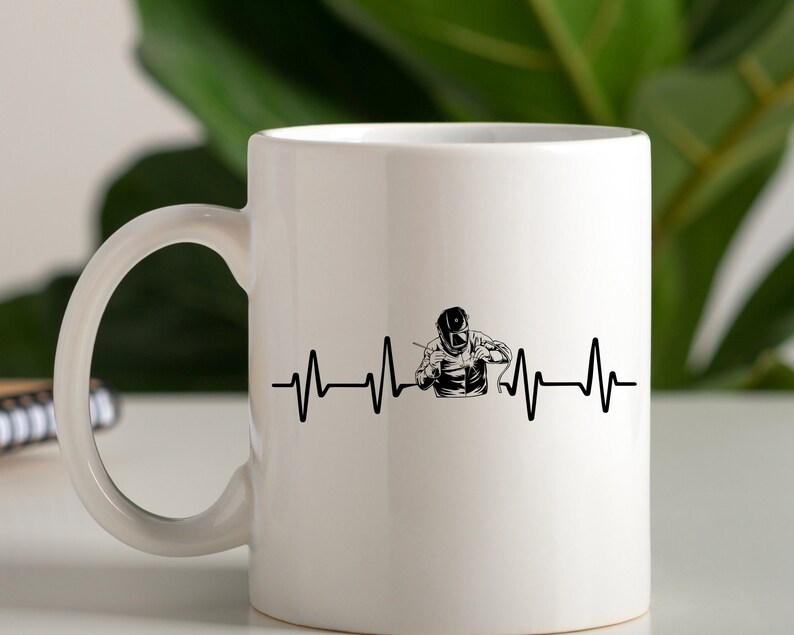 Puede incluir: Taza de cer&aacute;mica blanca con una silueta negra de un soldador con una m&aacute;scara de soldar y una l&iacute;nea de latido del coraz&oacute;n. El texto "My heart beats for welding" no es visible en la imagen.