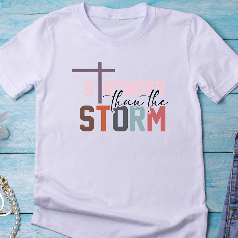 May include: White t-shirt with a colorful graphic design that says "Stronger than the storm" with a cross above the word "Stronger".