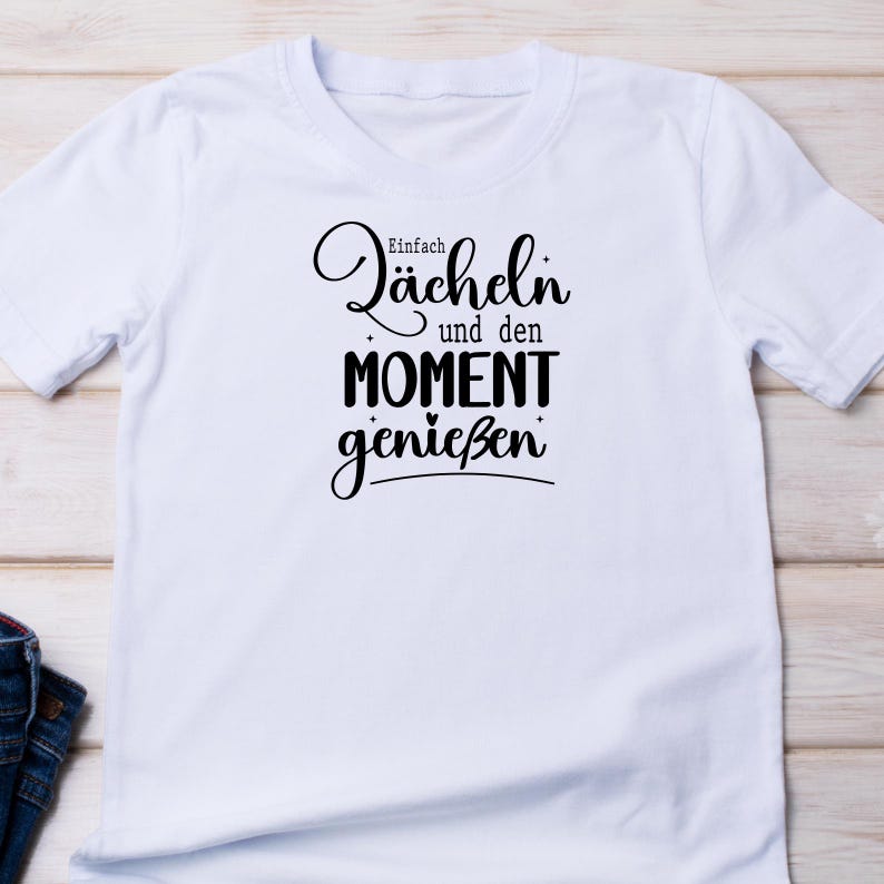 May include: White t-shirt with the German phrase "Einfach L&auml;cheln und den Moment genie&szlig;en" in black script. The text translates to "Simply smile and enjoy the moment". The shirt has short sleeves and a crew neck.