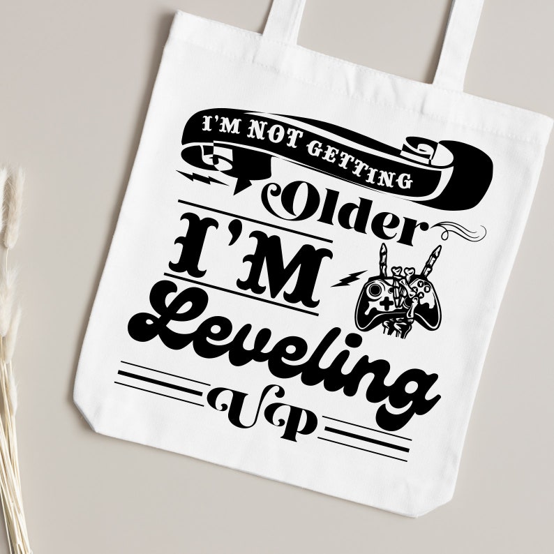 May include: White tote bag with a black graphic that says "I'm not getting older I'm leveling up" with a video game controller graphic.