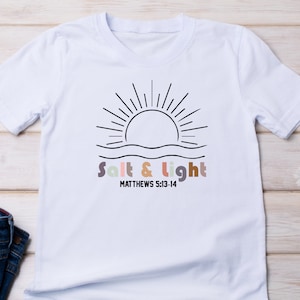 May include: White t-shirt with a black line drawing of a sun rising over water. The text "Salt & Light" is written in colorful letters below the sun, with the scripture reference "MATTHEWS 5:13-14" underneath.