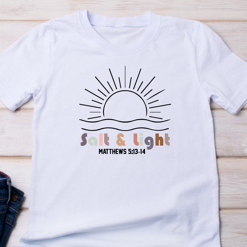 May include: White t-shirt with a black line drawing of a sun rising over water. The text "Salt & Light" is written in colorful letters below the sun, with the scripture reference "MATTHEWS 5:13-14" underneath.