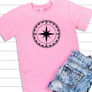 May include: A pink t-shirt with a black compass design on the front. The compass has a black star in the center and the letters N, S, E, and W around the edge.
