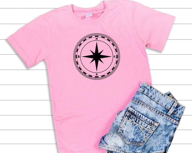 May include: A pink t-shirt with a black compass design on the front. The compass has a black star in the center and the letters N, S, E, and W around the edge.