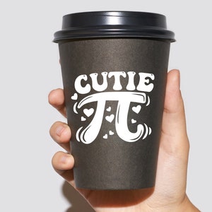 Puede incluir: Un vaso de caf&eacute; para llevar de color negro con un dise&ntilde;o blanco que dice "cutie pi" con corazones alrededor del s&iacute;mbolo pi.