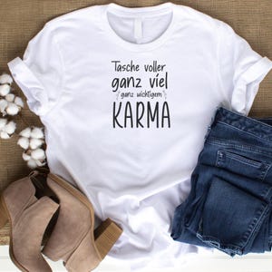 Pu&ograve; includere: T-shirt bianco con il testo tedesco "Tasche voller ganz viel ganz wichtigem KARMA" in nero. La maglietta &egrave; abbinata a jeans blu e stivaletti color cuoio. La maglietta &egrave; piegata e lo sfondo &egrave; di colore neutro.
