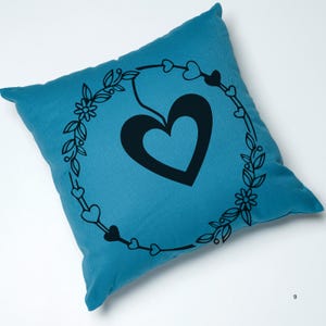 May include: A teal blue throw pillow with a black heart design inside a wreath of leaves and small hearts.