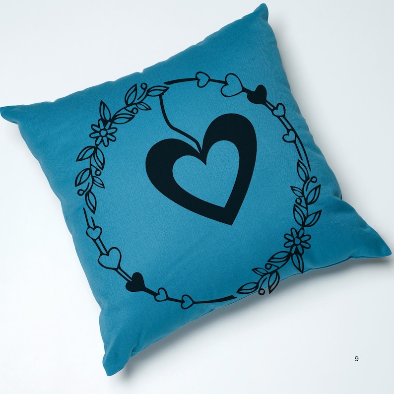 May include: A teal blue throw pillow with a black heart design inside a wreath of leaves and small hearts.