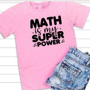 Puede incluir: Camiseta rosa con texto negro que dice "Math is my super power" con un s&iacute;mbolo de ra&iacute;z cuadrada antes y despu&eacute;s de la palabra "power".