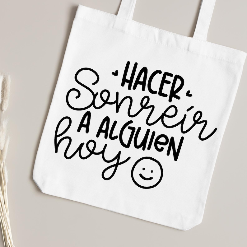 May include: White tote bag with black text that reads "Hacer sonre&iacute;r a alguien hoy" with a smiley face.