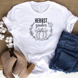 May include: White t-shirt with the text "HERBST zauber" in black, featuring a pumpkin and autumn leaves design. The shirt is paired with blue jeans and tan ankle boots, suggesting an autumn theme.