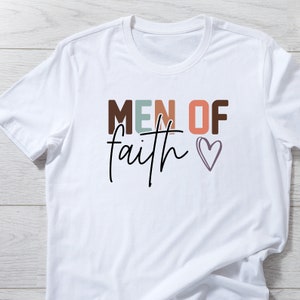 May include: White t-shirt with the text "MEN OF faith" in colorful letters and a small heart.
