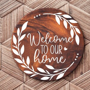 May include: A round wooden sign with a white wreath and the text "Welcome to our home" in a white cursive font.