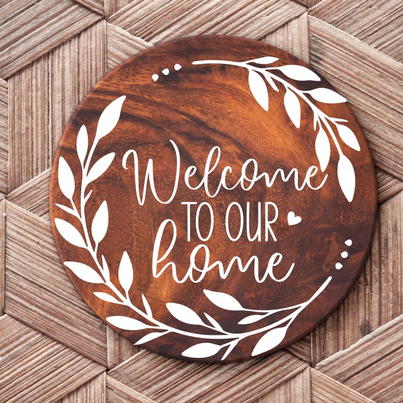 May include: A round wooden sign with a white wreath and the text "Welcome to our home" in a white cursive font.