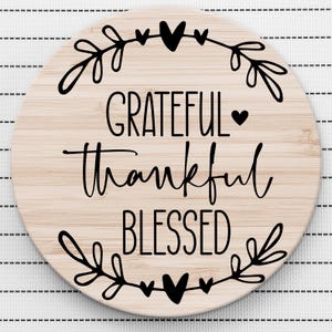 May include: Round wooden sign with the words "Grateful Thankful Blessed" in black script lettering. The sign is decorated with a wreath of leaves and hearts.