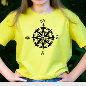 May include: A yellow t-shirt with a black compass design. The compass has the letters N, S, E, and W around it.