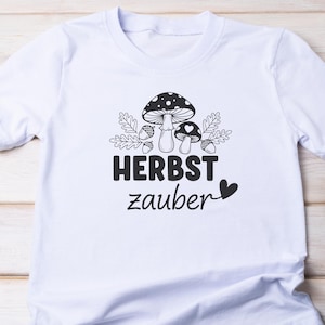 May include: White t-shirt with a black and white illustration of mushrooms, acorns, and leaves. The text "HERBST zauber" is printed below the graphic, with a heart symbol. The shirt has a crew neck.