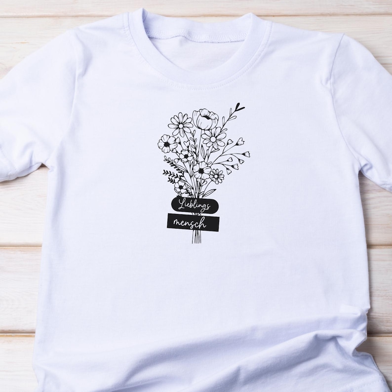 May include: White t-shirt featuring a black floral bouquet design with the words "Lieblingsmensch" in a stylized font. The bouquet includes various flowers and foliage, creating a delicate and artistic look. A casual and comfortable tee.