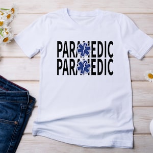 May include: White t-shirt with the words "PARAMEDIC" printed twice in black, with a blue star of life symbol between the words.