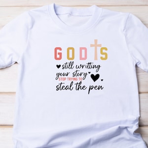 May include: White t-shirt with a black graphic that says "GOD'S still writing your story" with a cross and hearts. The text "STOP TRYING TO steal the pen" is also included.