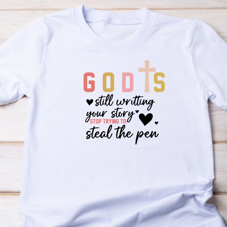 May include: White t-shirt with a black graphic that says "GOD'S still writing your story" with a cross and hearts. The text "STOP TRYING TO steal the pen" is also included.