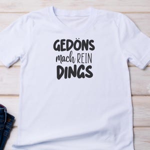 Pu&ograve; includere: T-shirt bianca con la scritta nera "GED&Ouml;NS mach REIN DINGS". La maglietta a maniche corte ha un girocollo e il testo &egrave; centrato. La maglietta &egrave; realizzata in un materiale morbido.