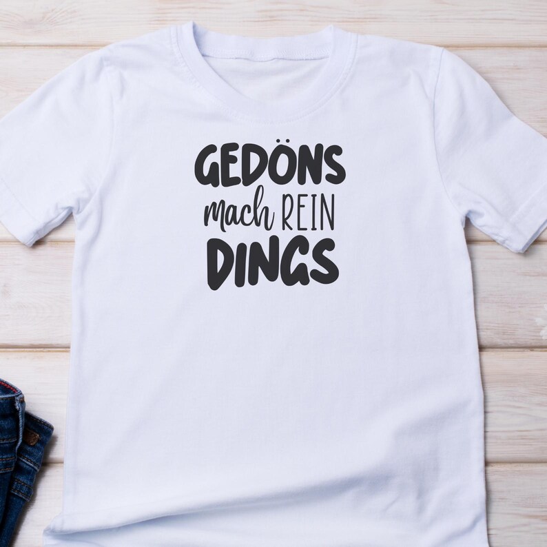 Pu&ograve; includere: T-shirt bianca con la scritta nera "GED&Ouml;NS mach REIN DINGS". La maglietta a maniche corte ha un girocollo e il testo &egrave; centrato. La maglietta &egrave; realizzata in un materiale morbido.