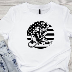 Puede incluir: Camiseta blanca con un gr&aacute;fico en blanco y negro de un soldador con una m&aacute;scara de soldar y sosteniendo una antorcha de soldar. El gr&aacute;fico est&aacute; sobre un c&iacute;rculo con un dise&ntilde;o de bandera estadounidense en blanco y negro.