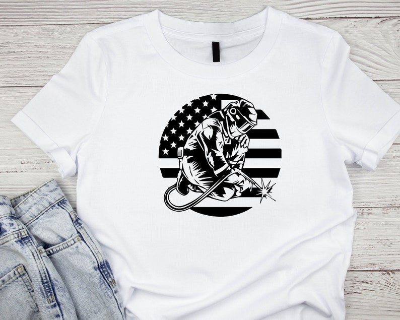 Puede incluir: Camiseta blanca con un gr&aacute;fico en blanco y negro de un soldador con una m&aacute;scara de soldar y sosteniendo una antorcha de soldar. El gr&aacute;fico est&aacute; sobre un c&iacute;rculo con un dise&ntilde;o de bandera estadounidense en blanco y negro.