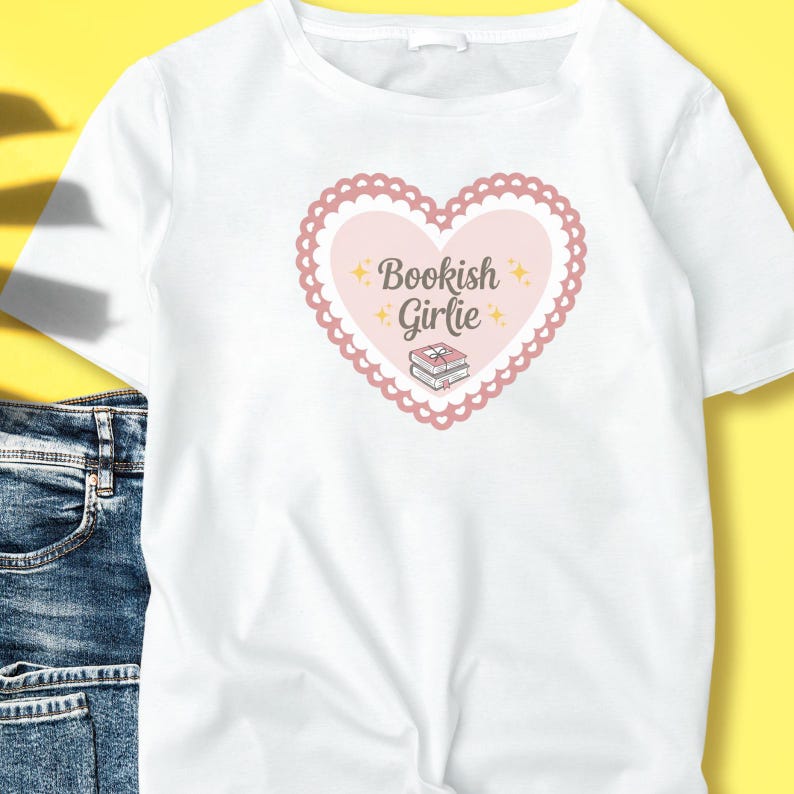 K&ouml;nnte beinhalten: Wei&szlig;es T-Shirt mit einem rosa Herz-Design, das die Worte "Bookish Girlie" und einen B&uuml;cherstapel enth&auml;lt. Ein weiches, feminines Design, ideal f&uuml;r B&uuml;cherliebhaber.