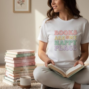 K&ouml;nnte beinhalten: Wei&szlig;es T-Shirt mit dem Text "BOOKS ARE MY HAPPY PLACE" in Pastellfarben. Ein Stapel B&uuml;cher in verschiedenen Farben und eine Tasse befinden sich im Vordergrund. Die Person h&auml;lt ein offenes Buch.