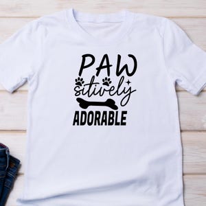 Puede incluir: Camiseta blanca con texto negro que dice "Paw-sitively Adorable" con huellas de patas y un gr&aacute;fico de hueso.