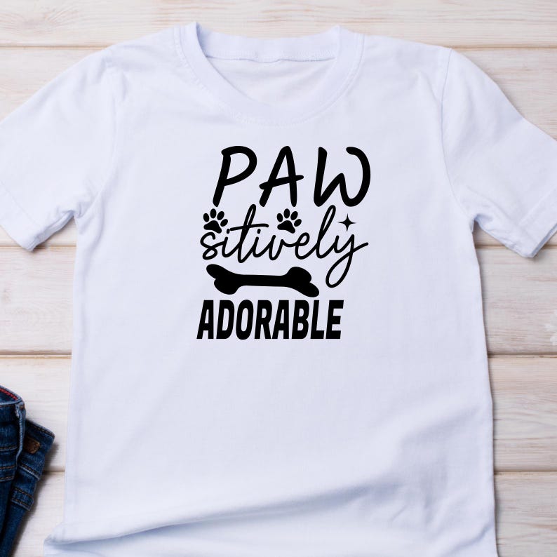 Puede incluir: Camiseta blanca con texto negro que dice "Paw-sitively Adorable" con huellas de patas y un gr&aacute;fico de hueso.