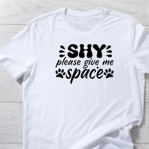 Puede incluir: Camiseta blanca con texto negro que dice "SHY please give me space" con huellas de patas a cada lado de la palabra "space".