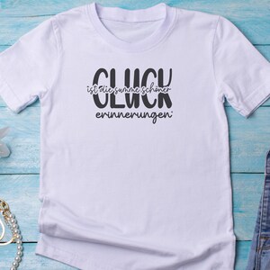 May include: White t-shirt with the German text "GL&Uuml;CK ist die summe sch&ouml;ner erinnerungen" in black. The shirt has short sleeves and a crew neck. The text translates to "Happiness is the sum of beautiful memories."