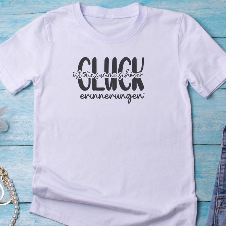 May include: White t-shirt with the German text "GL&Uuml;CK ist die summe sch&ouml;ner erinnerungen" in black. The shirt has short sleeves and a crew neck. The text translates to "Happiness is the sum of beautiful memories."