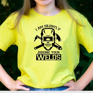 Puede incluir: Una camiseta amarilla con un gr&aacute;fico negro de una m&aacute;scara de soldador y el texto "Estoy juzgando tus soldaduras en silencio".