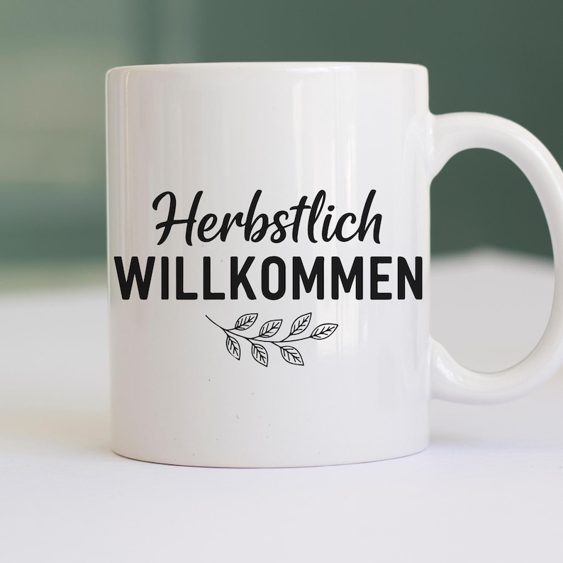 May include: White ceramic mug featuring the German words "Herbstlich WILLKOMMEN" in black script and block letters, with a leaf design. The mug has a curved handle and is suitable for hot drinks.