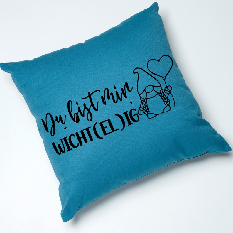 May include: Teal square pillow with black text and a gnome illustration. The text reads "Du bist min WICHT(EL)IG". The gnome is holding a heart-shaped balloon. The pillow is a solid teal color.